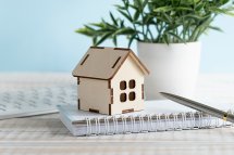 © Inna / stock.adobe.com Miniaturhaus aus Holz auf Schreibblock vor Topfpflanze